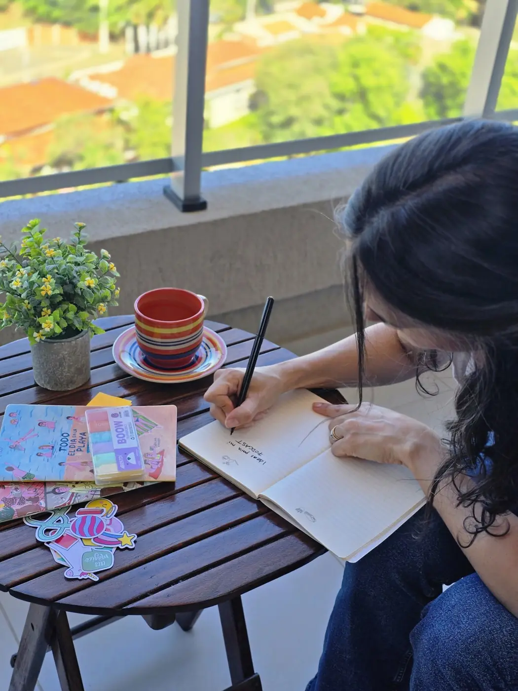 Aura escribe en un cuaderno en el balcón de Gánica Estudio, con café y libros para colorear