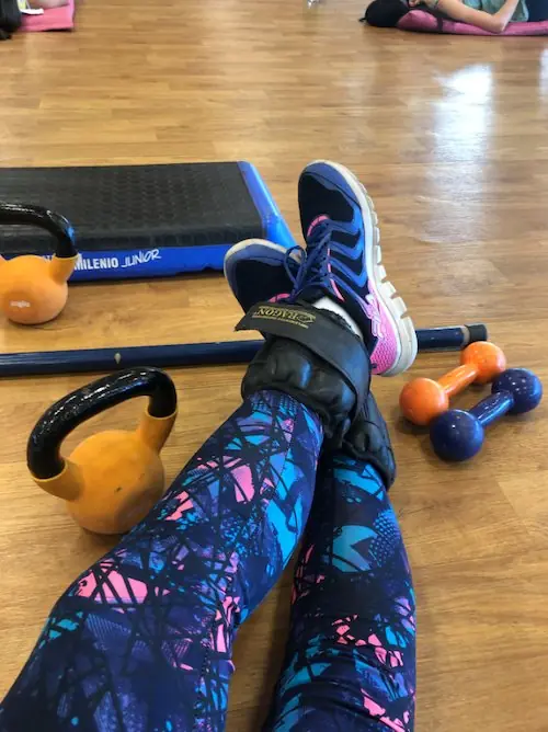 Piernas con calzas estampadas apoyadas en un step, junto a pesas y una pesa rusa en el piso del gimnasio